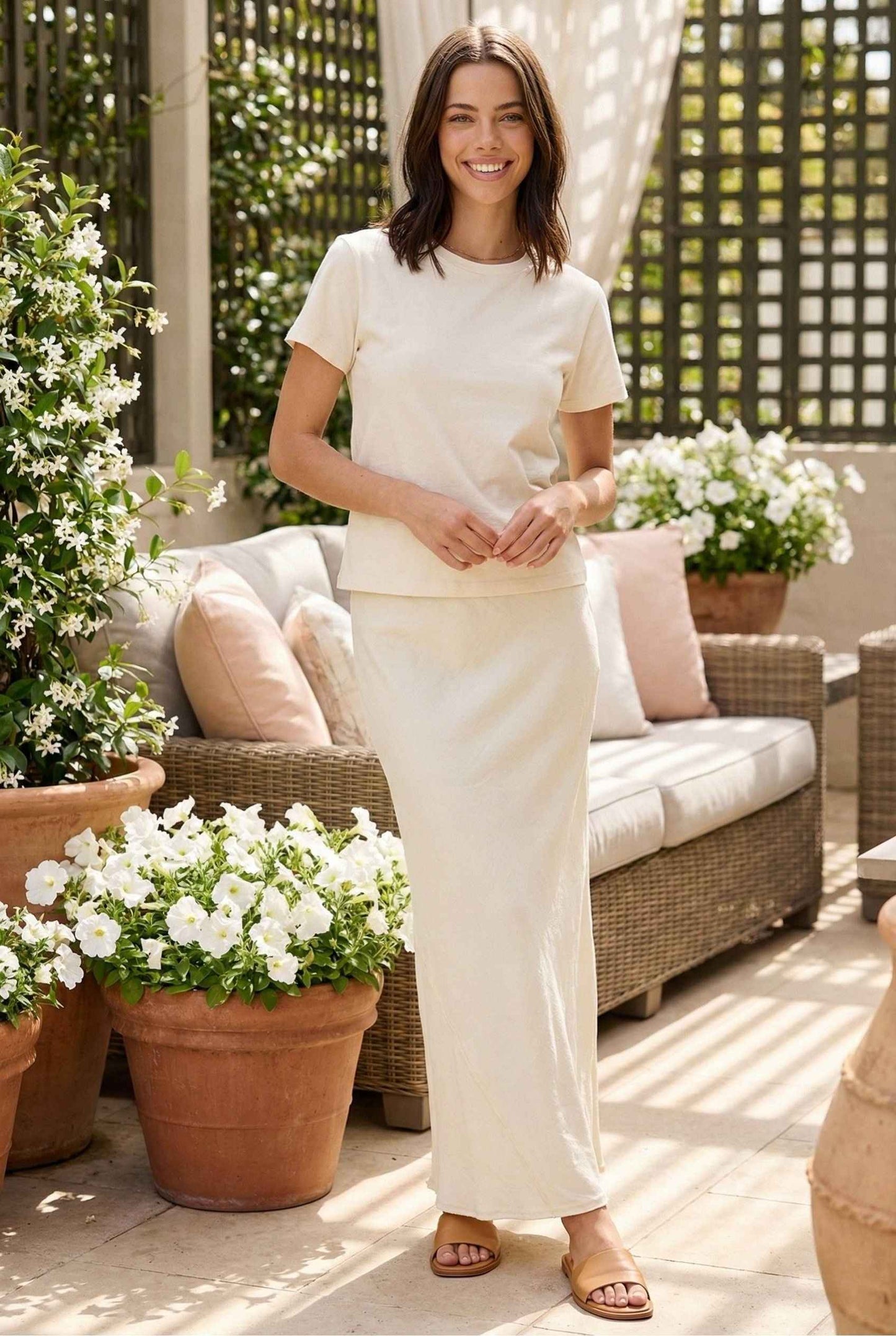 Model wearing Veda Skirt in Cream Satin Drawstring Maxi Skirt paired with basic t-shirt and leather slides posing on patio