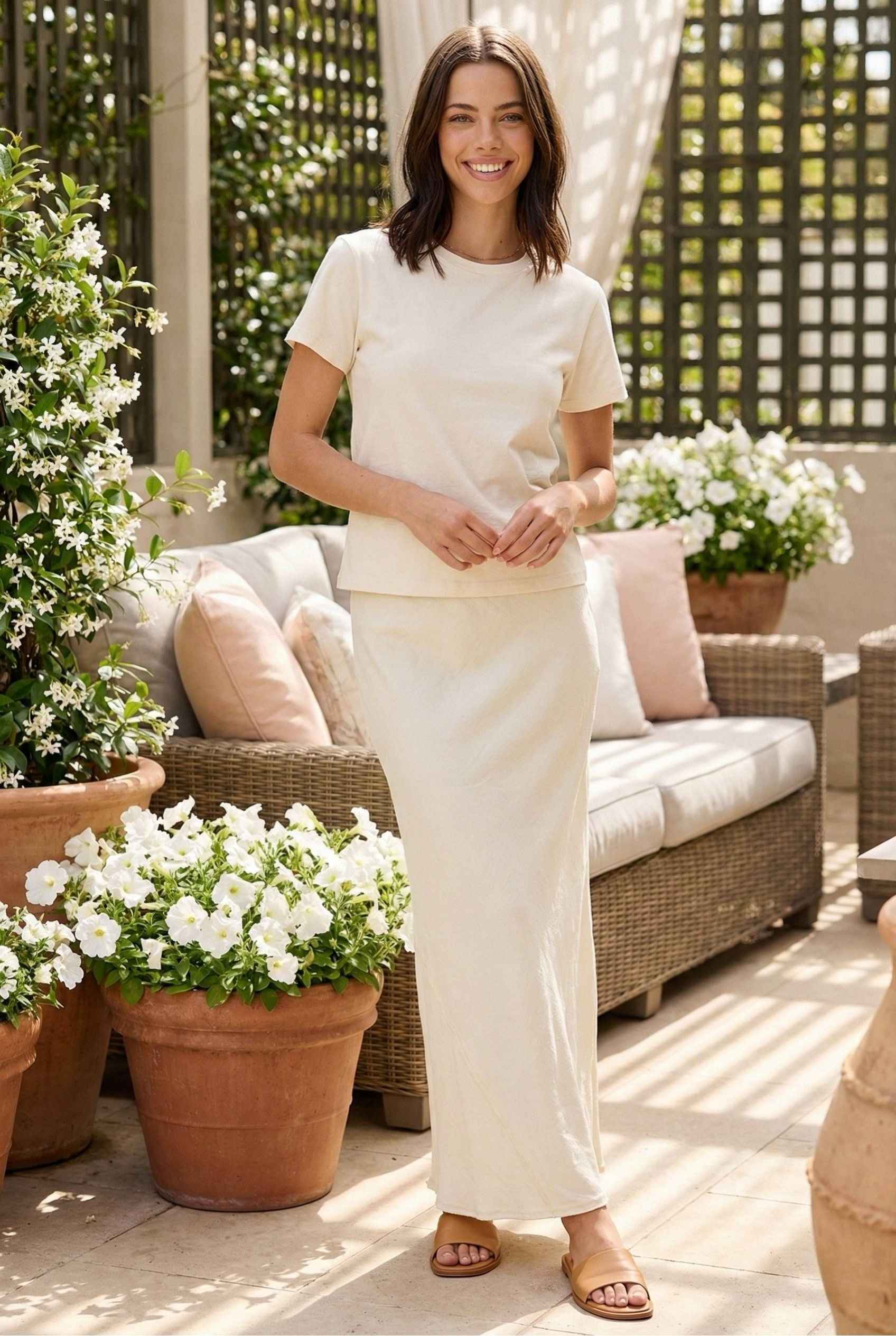 Model wearing Veda Skirt in Cream Satin Drawstring Maxi Skirt paired with basic t-shirt and leather slides posing on patio