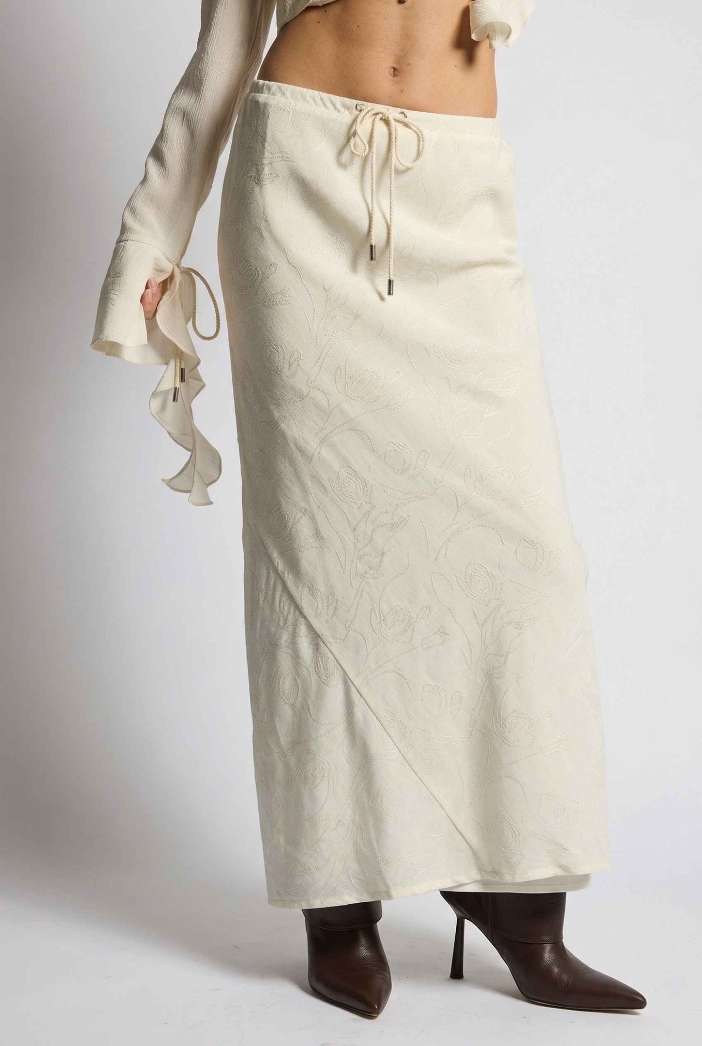 Model wearing Veda Skirt in Cream Satin Drawstring Maxi Skirt paired with matching ruffle tie bouse and booties
