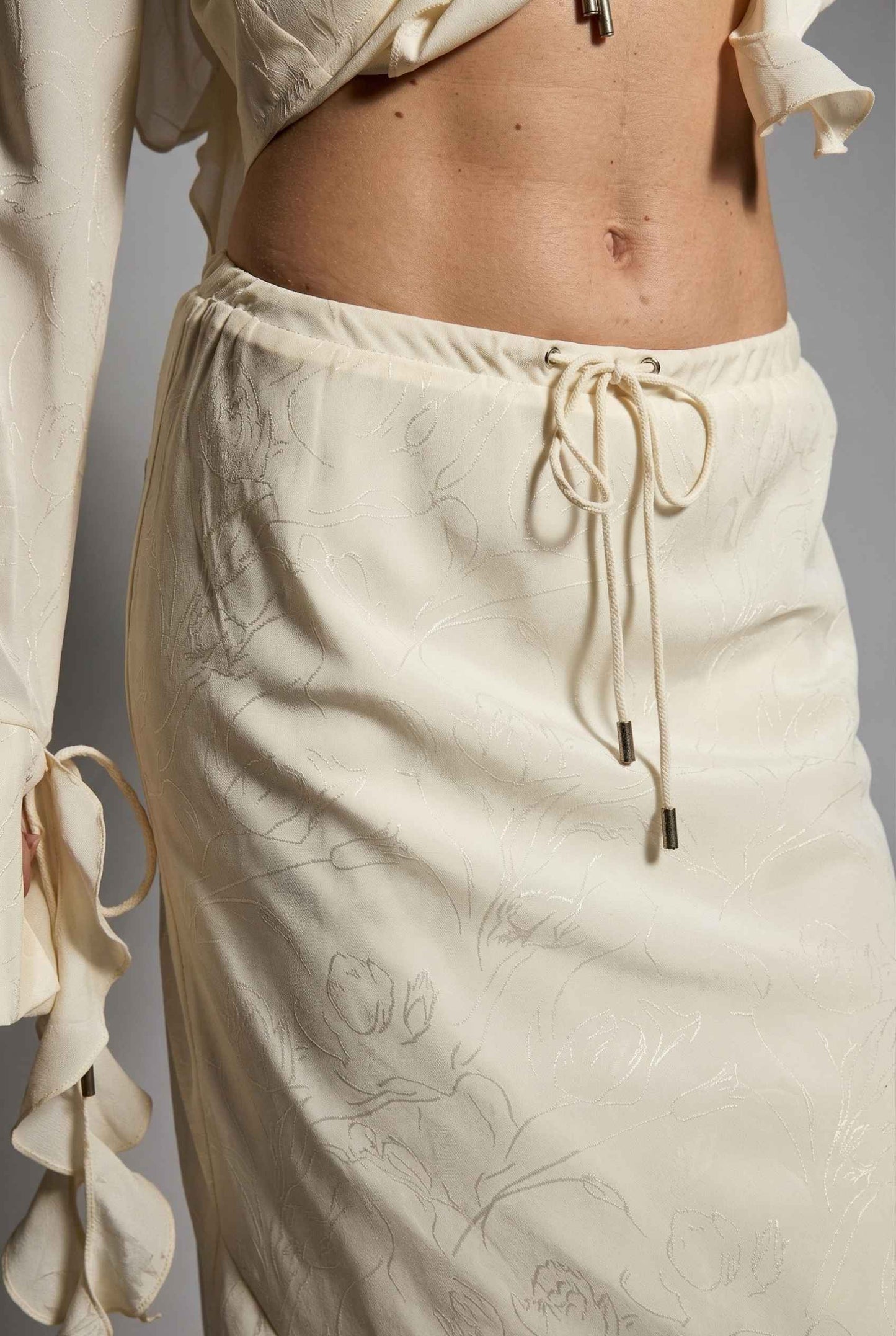 Model wearing Veda Skirt in Cream Satin Drawstring Maxi Skirt paired with matching ruffle tie bouse close up tie up detail