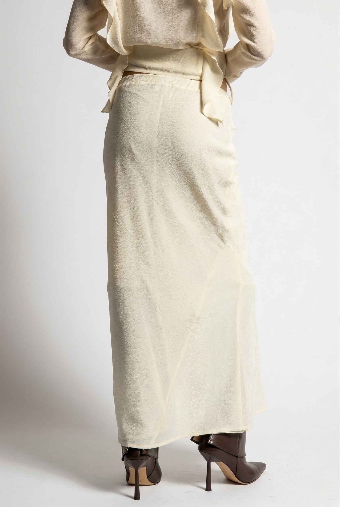 Model wearing Veda Skirt in Cream Satin Drawstring Maxi Skirt paired with matching ruffle tie bouse and booties