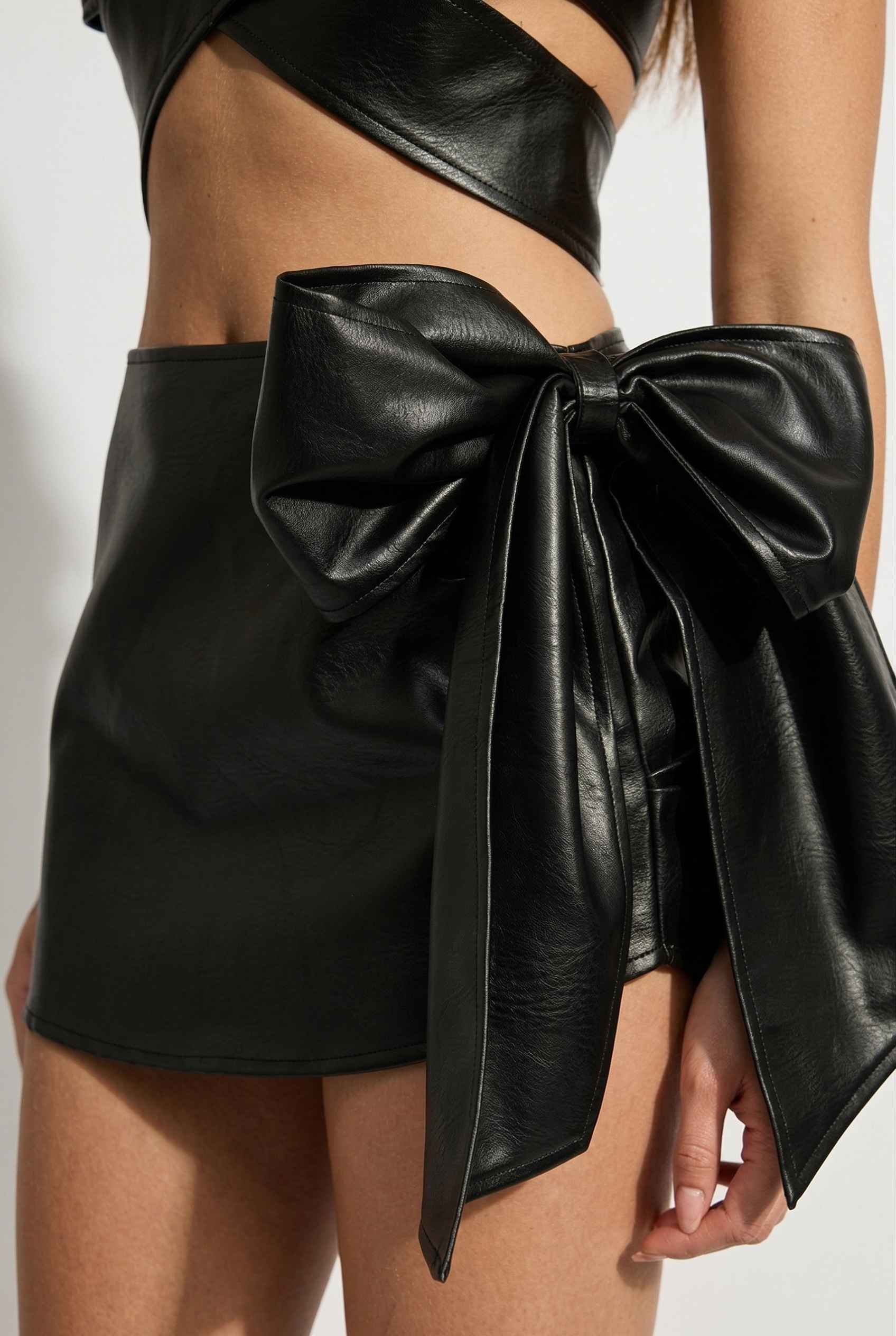 Model wearing Vesper Skirt in Black Vegan Leather Mini Skirt with Statement Bow paired with matching bustier top up close giant bow detail