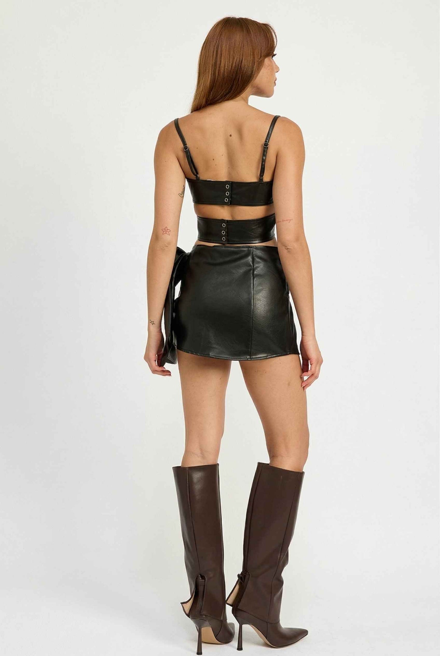 Model wearing Vesper Skirt in Black Vegan Leather Mini Skirt with Statement Bow paired with matching bustier top and knee-high boots