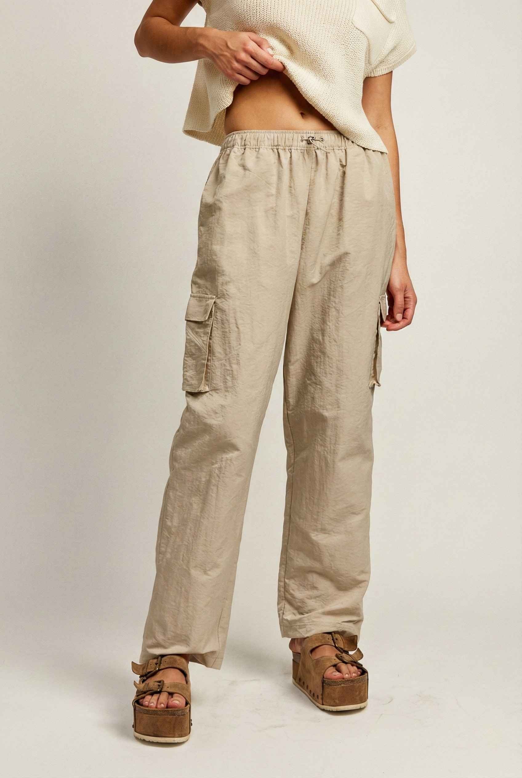Model wearing Wren Pants 100% Nylon Parachute Cargo Pants in Taupe with t-shirt and platform sandals on plain background