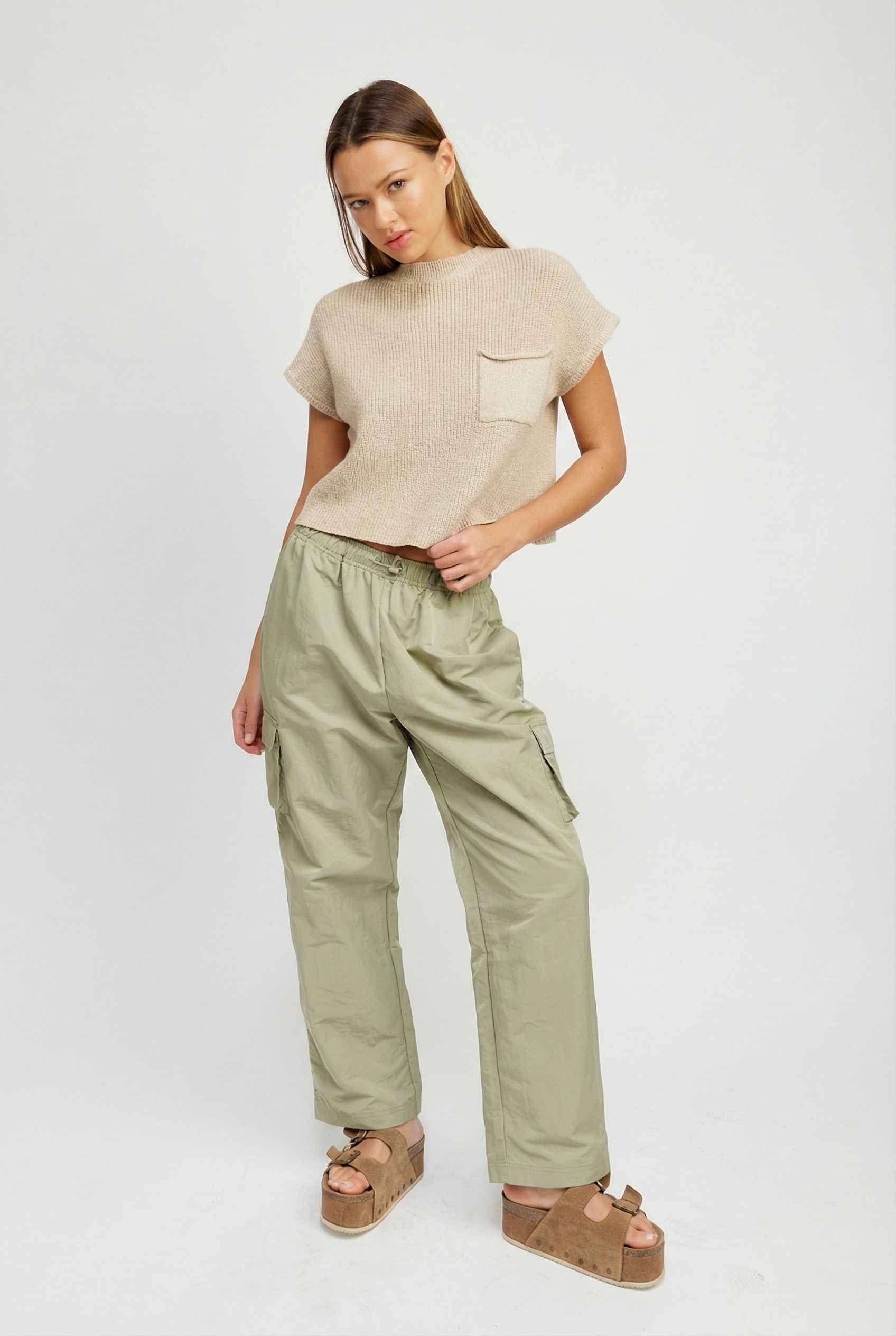 Model wearing Wren Pants 100% Nylon Parachute Cargo Pants in Olive with t-shirt and platform sandals on plain background