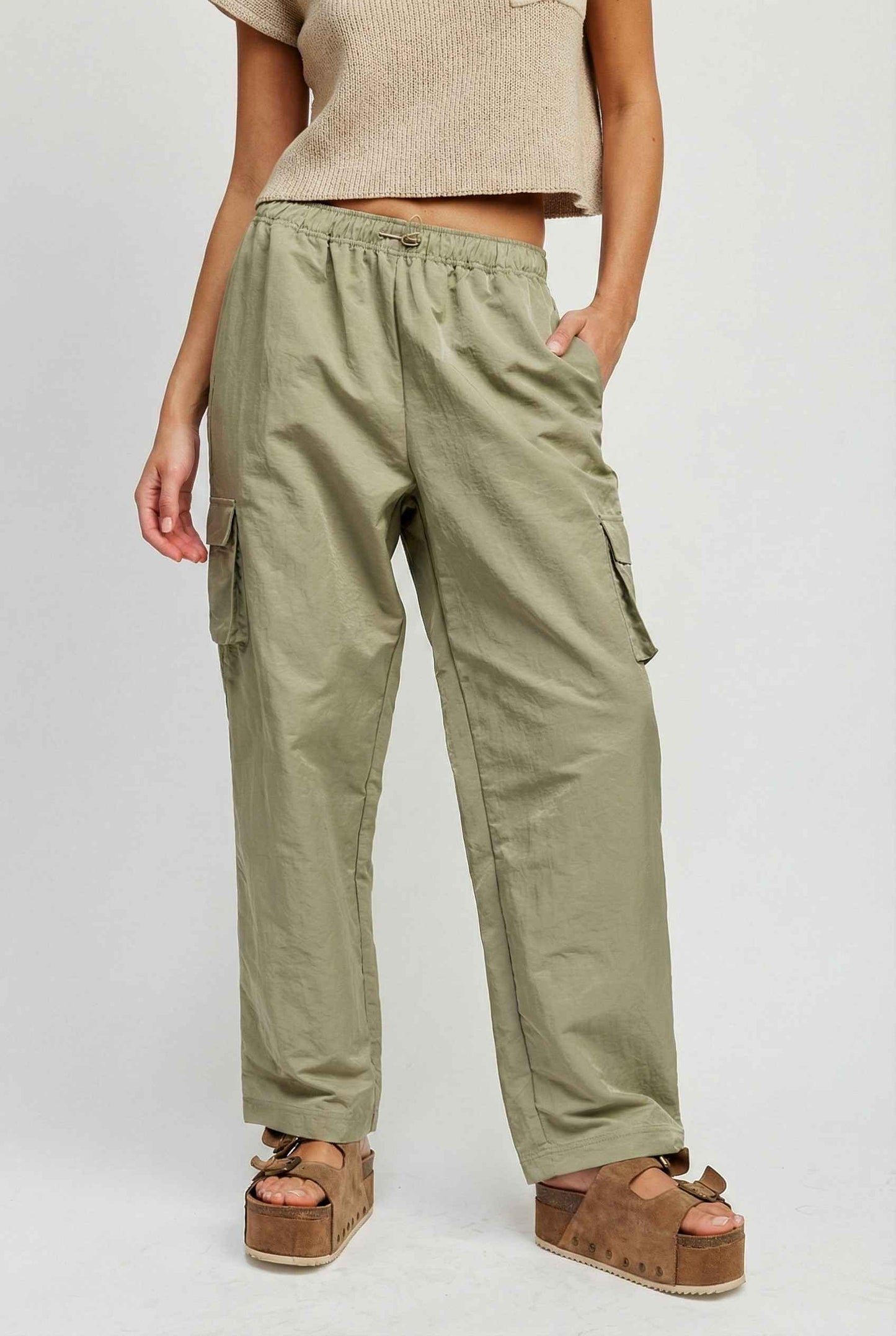 Model wearing Wren Pants 100% Nylon Parachute Cargo Pants in Olive with t-shirt and platform sandals on plain background
