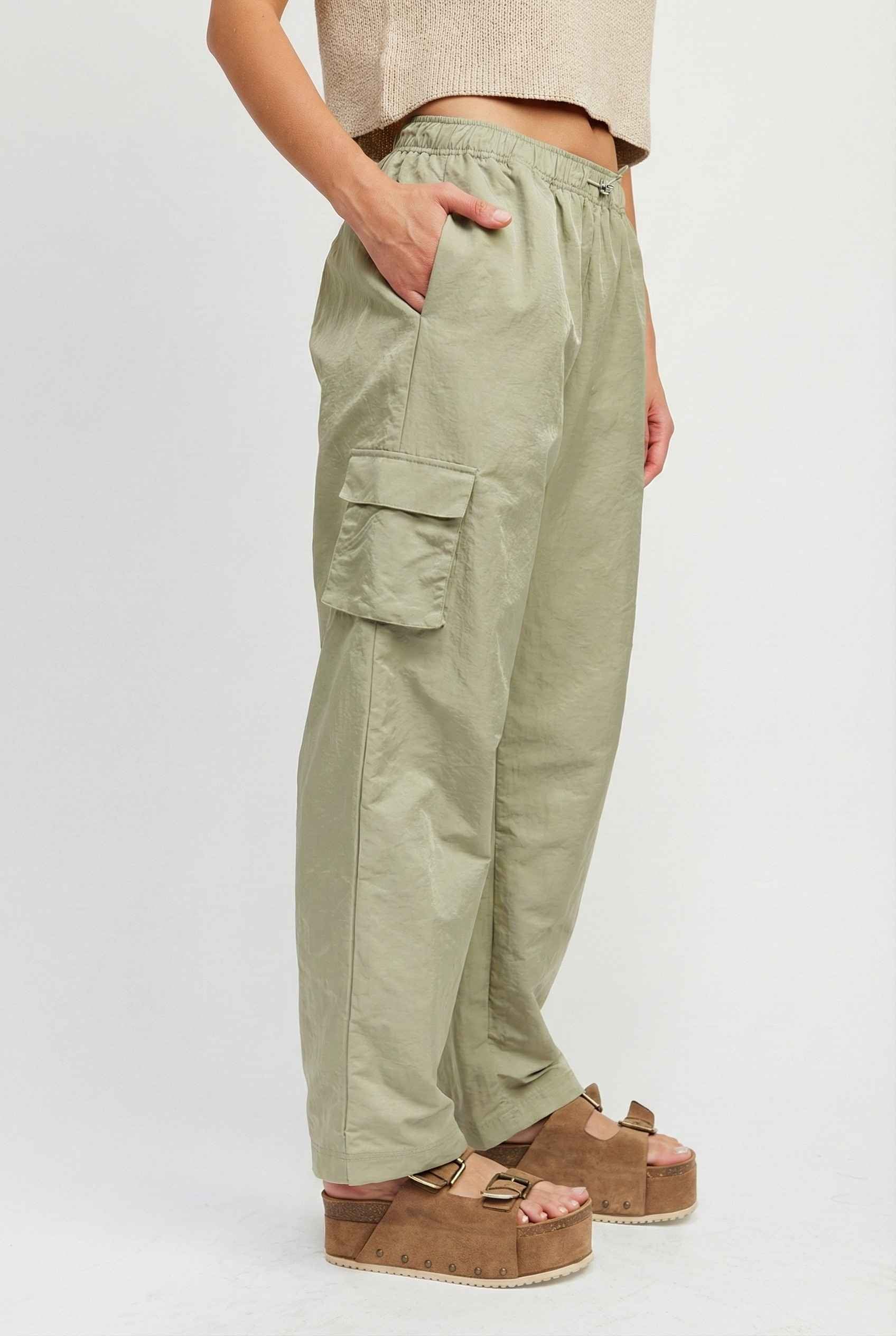 Model wearing Wren Pants 100% Nylon Parachute Cargo Pants in Olive with t-shirt and platform sandals on plain background