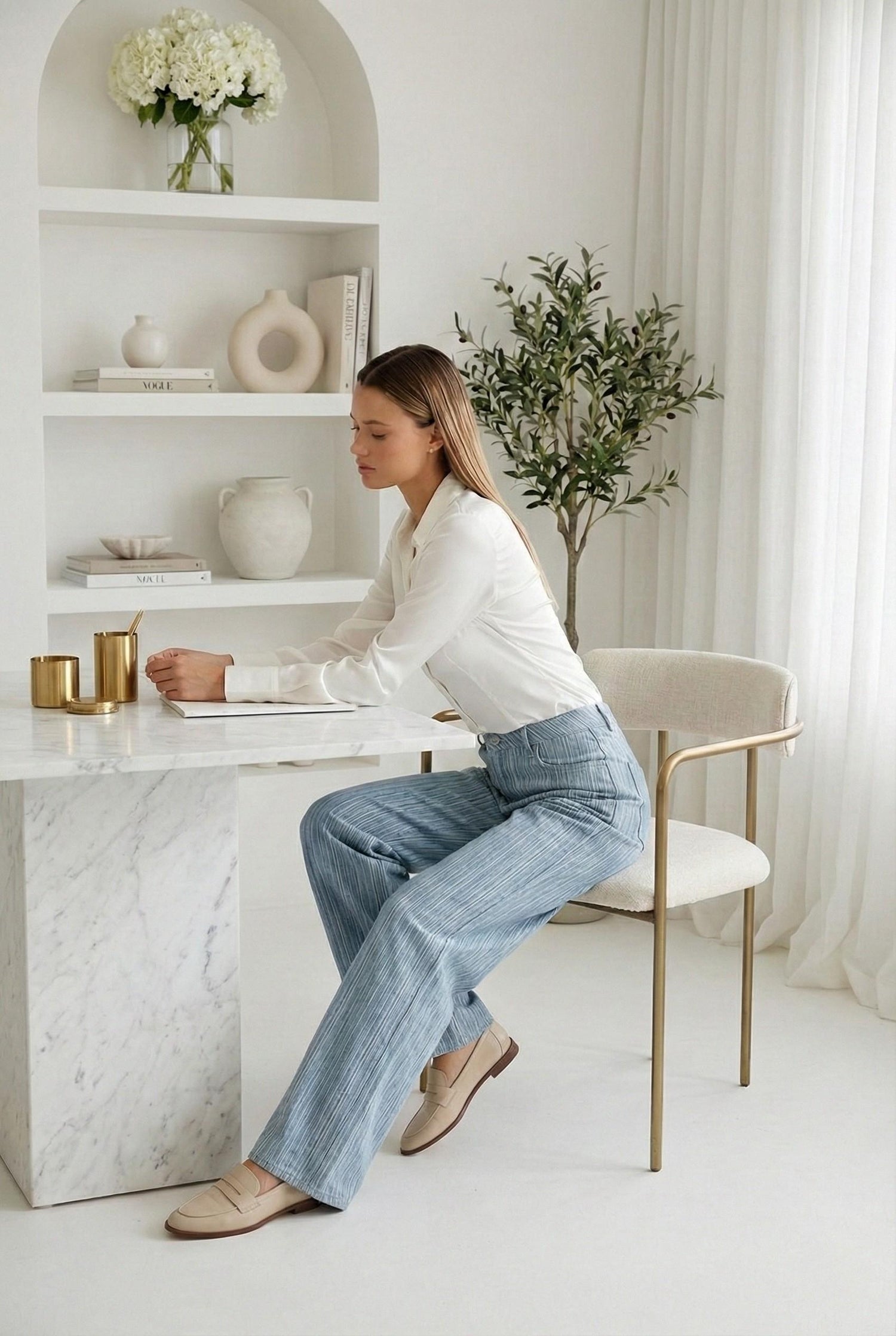 Model wearing Emory Park Mid Rise Wide Leg Pants in blue Denim pinstripe with blouse and loafers sitting in luxury minimalist office
