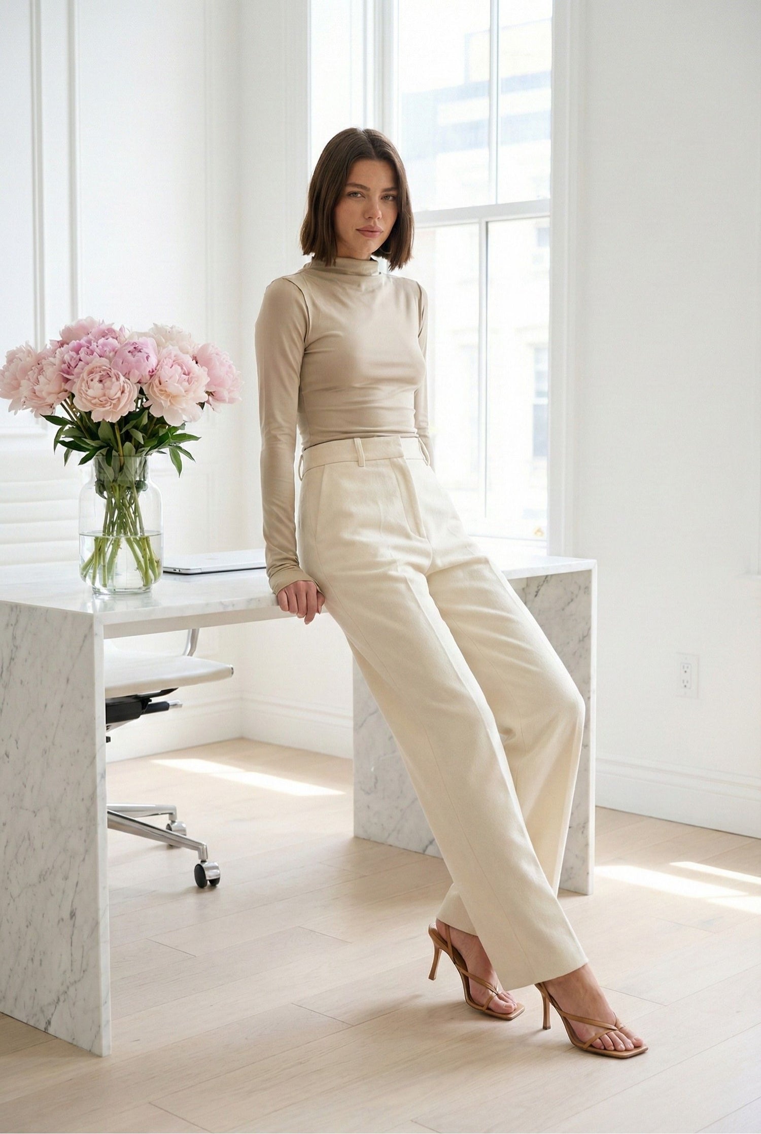 Model wearing Emory Park Mock Neck Long Sleeve Jersey Top in Latte with trousers and heels posing in minimalist office with a vase of flowers