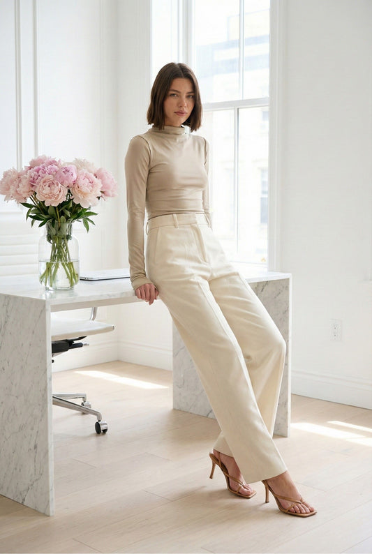 Model wearing Emory Park Mock Neck Long Sleeve Jersey Top in Latte with trousers and heels posing in minimalist office with a vase of flowers